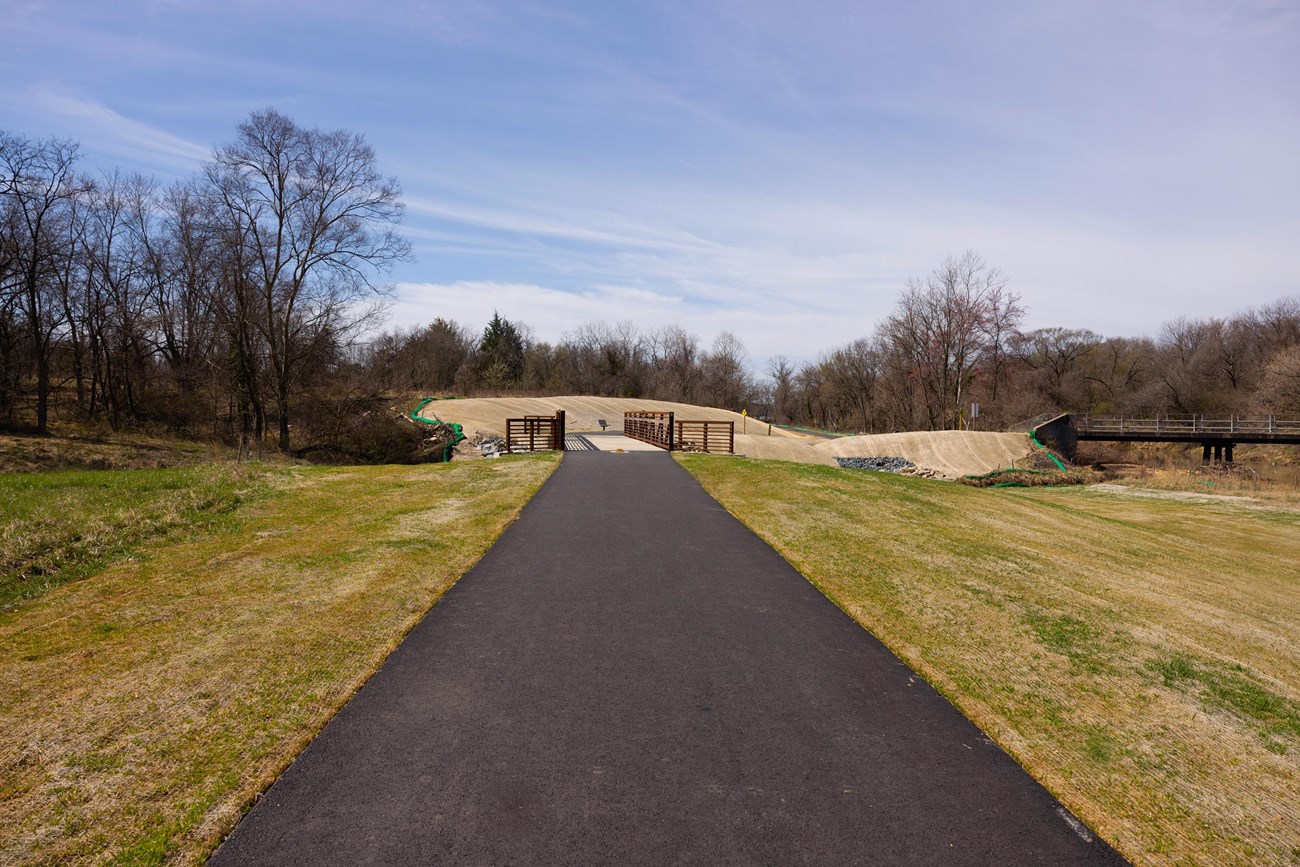 Newly paved trail leading to a new bridge. Grass is seen on both sides of the newly paved path. Trees are seen around the bridge. The bridge is concrete with red, open siding.