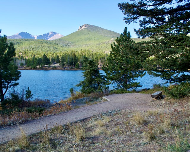 Trail to an overlook with a bench along a lake with mountains.