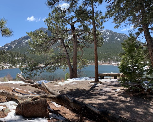 Dirt trail with a little snow overlooking a lake and mountains.