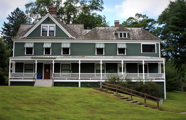 Green two-and-a-half story house with white trim, wraparound porch, wood roof, and striped awnings.