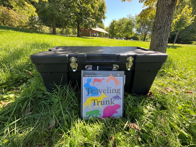 black plastic trunk with metal clasps sitting in the grass with a colorful binder in front.