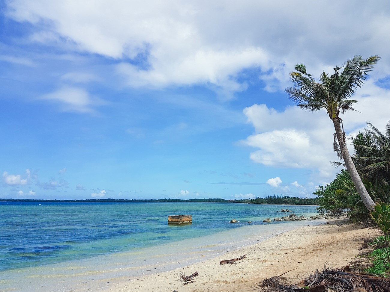 A sandy beach and clear blue water. A concrete block is in the water.
