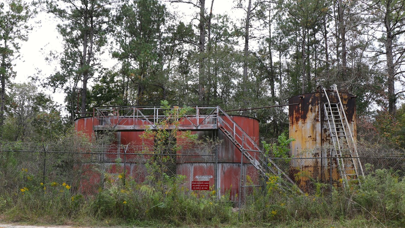 2 large red rusty tanks and 1 large yellow rusty tank with a ladder surrounded by a chainlink fence and forest.