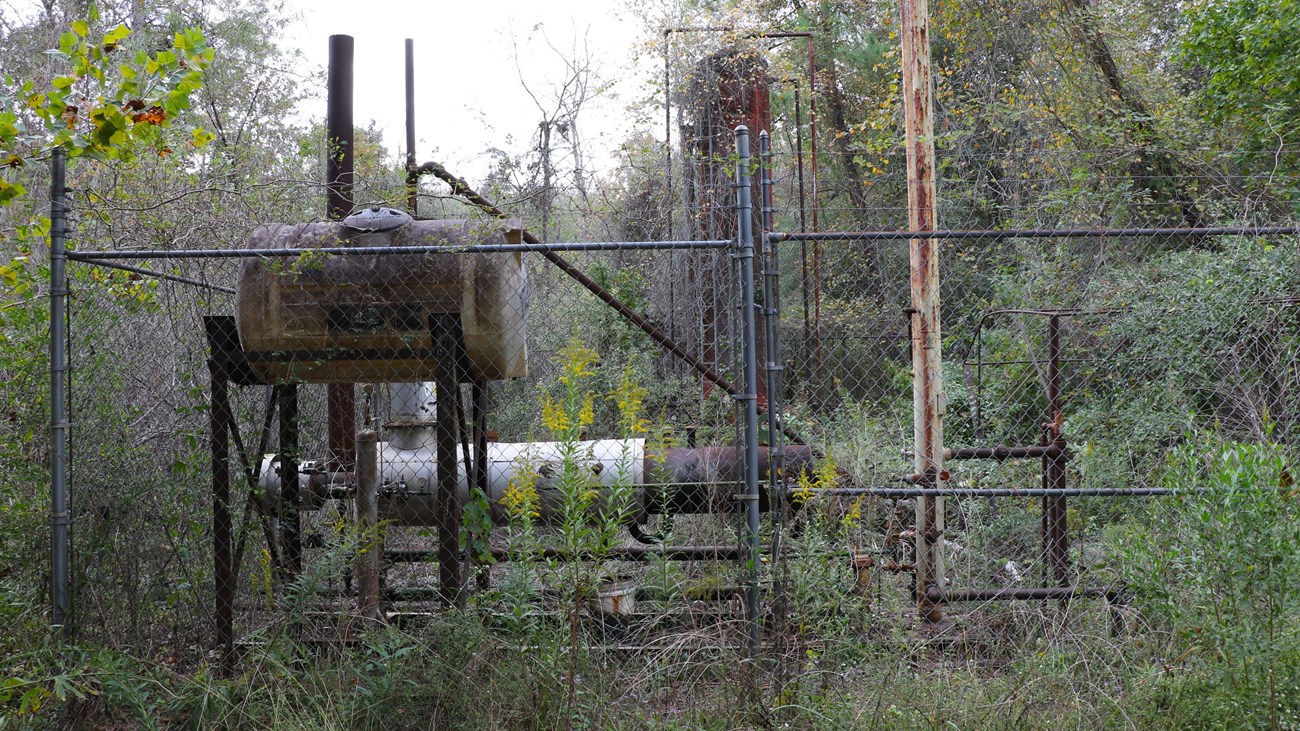 abandoned oil machinery and tanks surrounded by a chainlink fence and overgrown vegetation