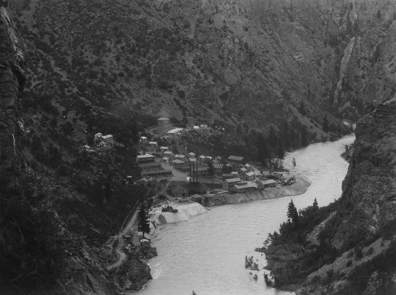 Historic black and white image of a town built alongside a river. Canyon walls rise on either side of the town. Buildings rise up the canyon walls and a power plant is right next to the river.