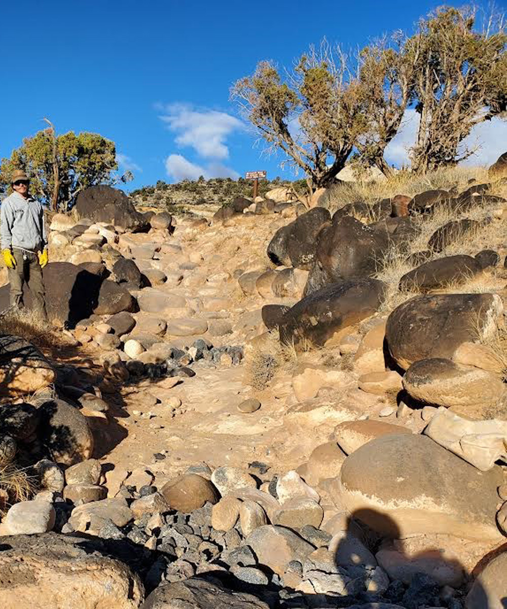 Stone-lined trail with uneven surfaces and missing steps, surrounded by boulders and desert vegetation under a bright blue sky