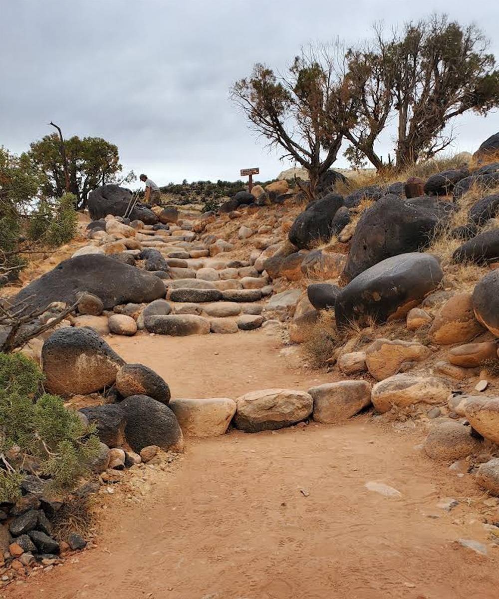 Stone-lined trail with uneven surfaces and missing steps, surrounded by boulders and desert vegetation under a bright blue sky