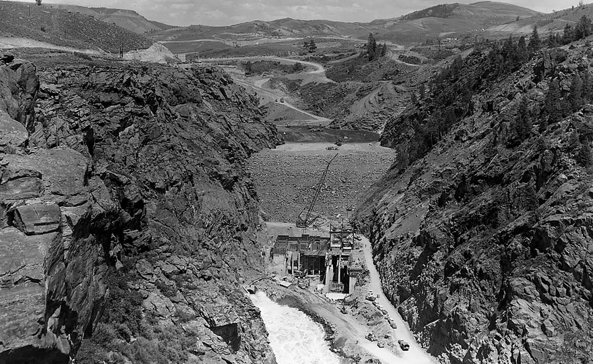 Historic black and white image of an earthen dam construction between two canyon walls.