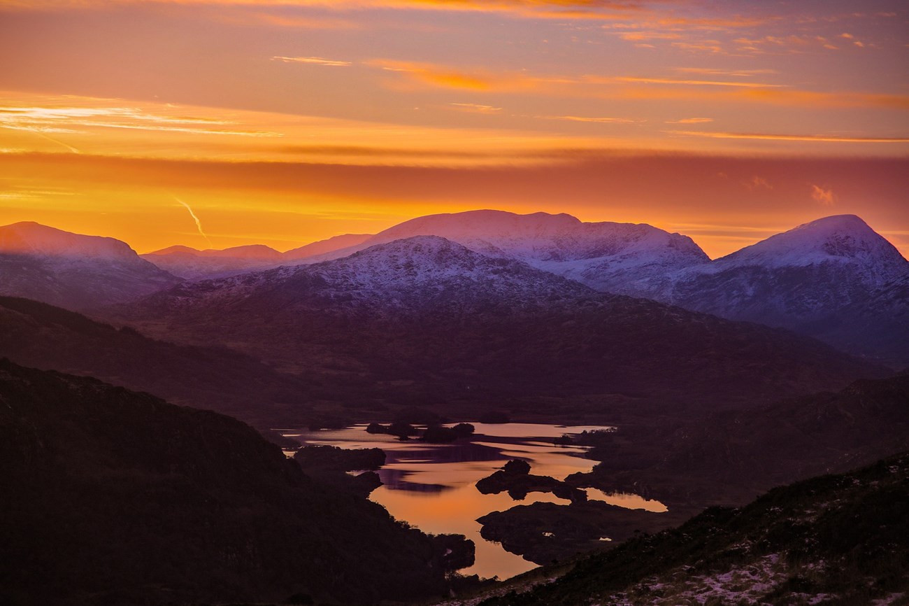 A range of mountains colored purple from the sun setting are seen in the distance. The reflection of an orange sky is seen in the lake at its base.