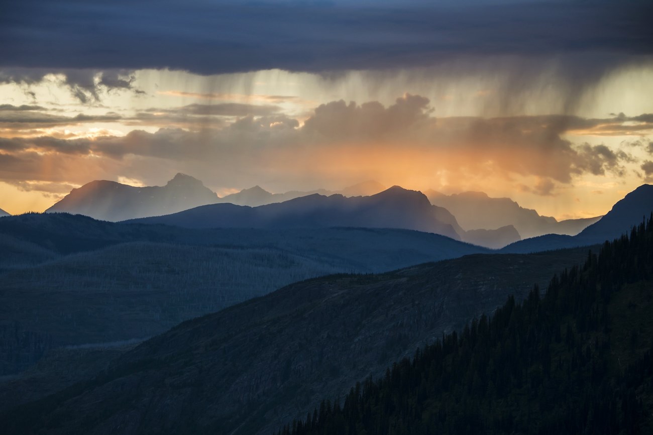 A range of mountains colored purple from the sun setting are seen in the distance. The reflection of an orange sky is seen in the lake at its base.