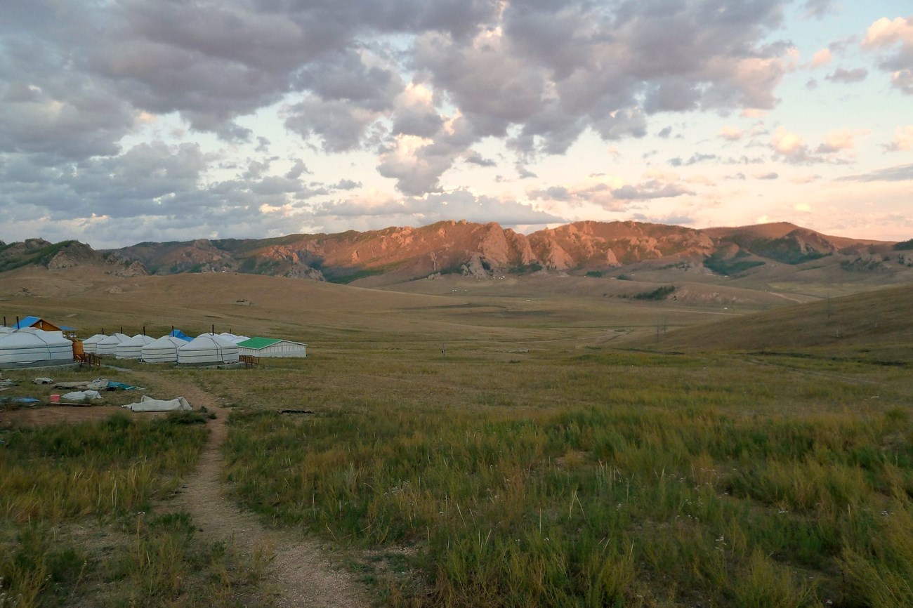 Wide open plains and hills with green and yellow grasses lead up to a wall of mountains lit by the sun setting. A camp of white tents at the end of a dirt path is seen on the left side.