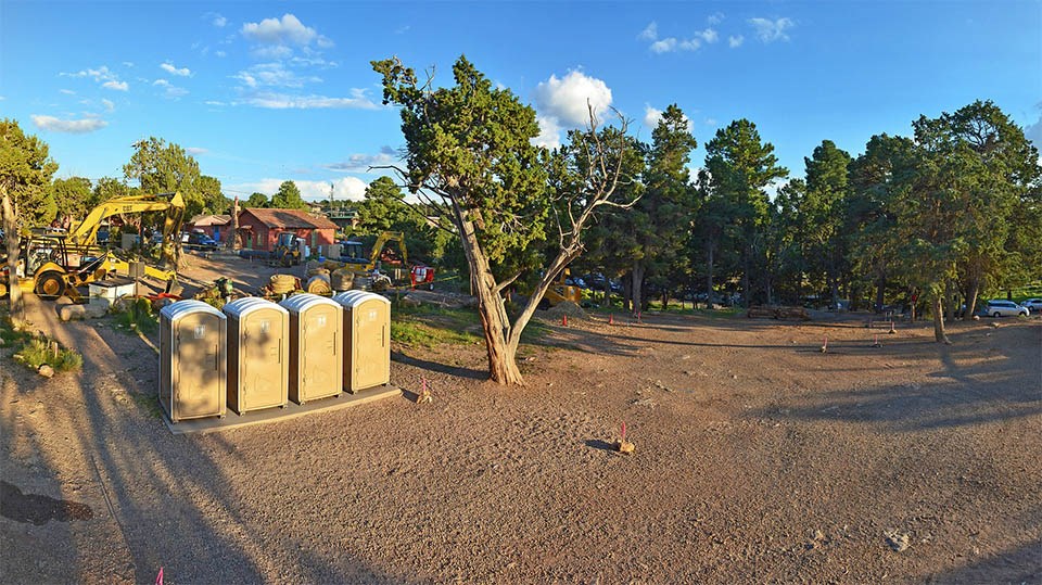 An undeveloped area next to the Bright Angel Trailhead shows the ground, trees, and four Porta-a-potties.