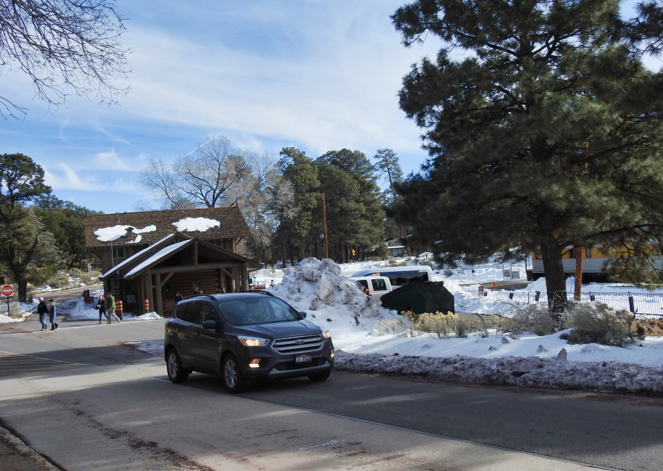 People, train, and cars next to railroad depot building, trees in background