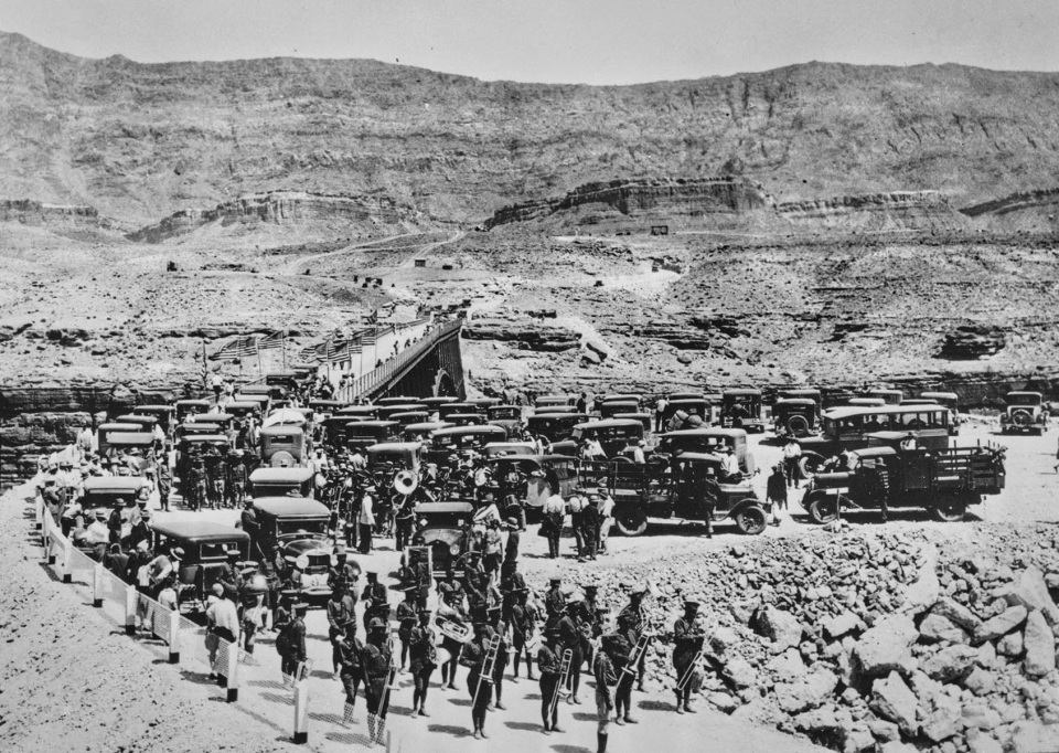 Large group of people with cars, bridge and hills in background