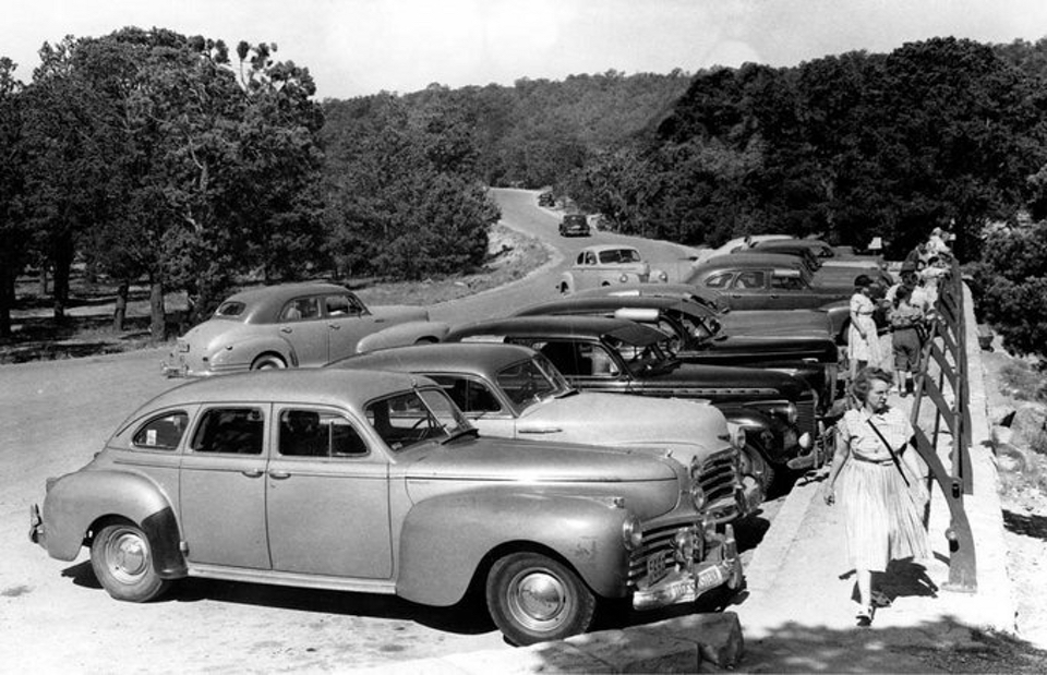 Multiple parked cars, people, a fence, road and trees
