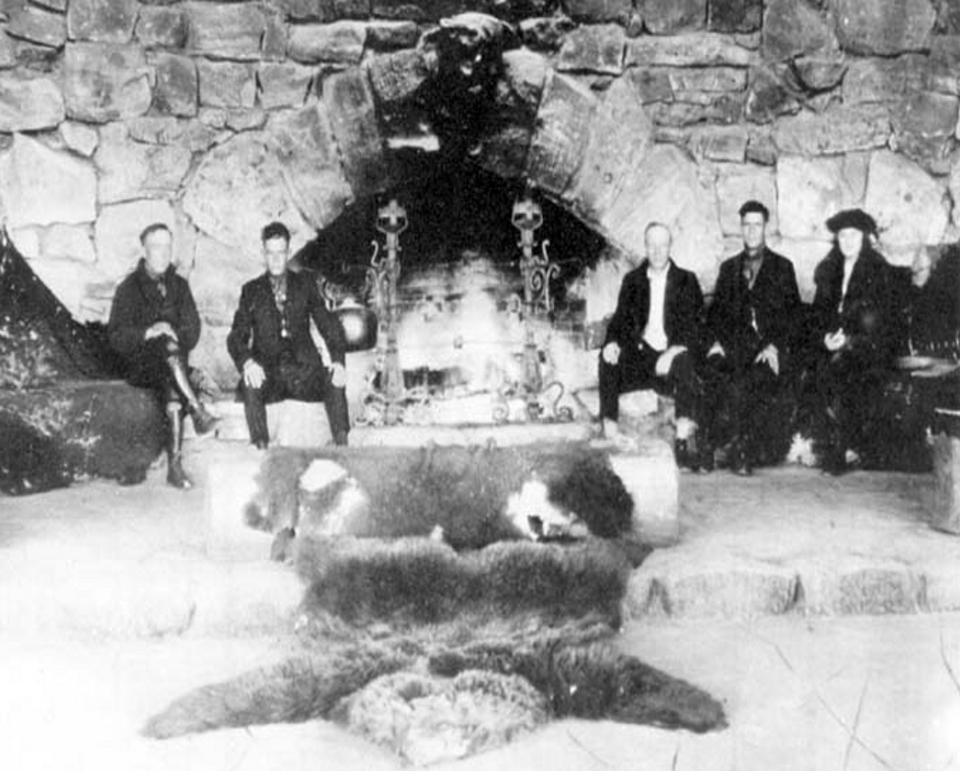 Men sitting in front of stone fireplace with candles and bear rug on floor