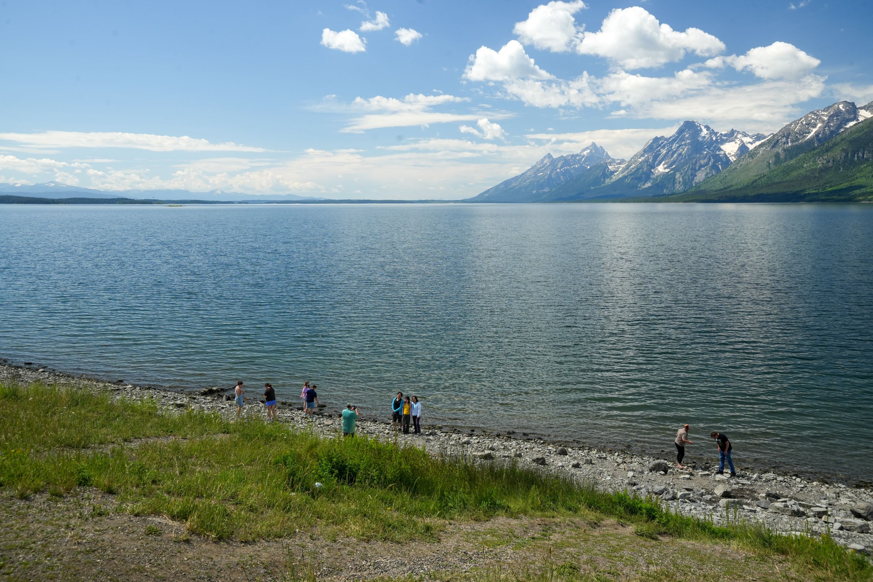 Visitors stand near the edge of a lake with the teton range in the distance.