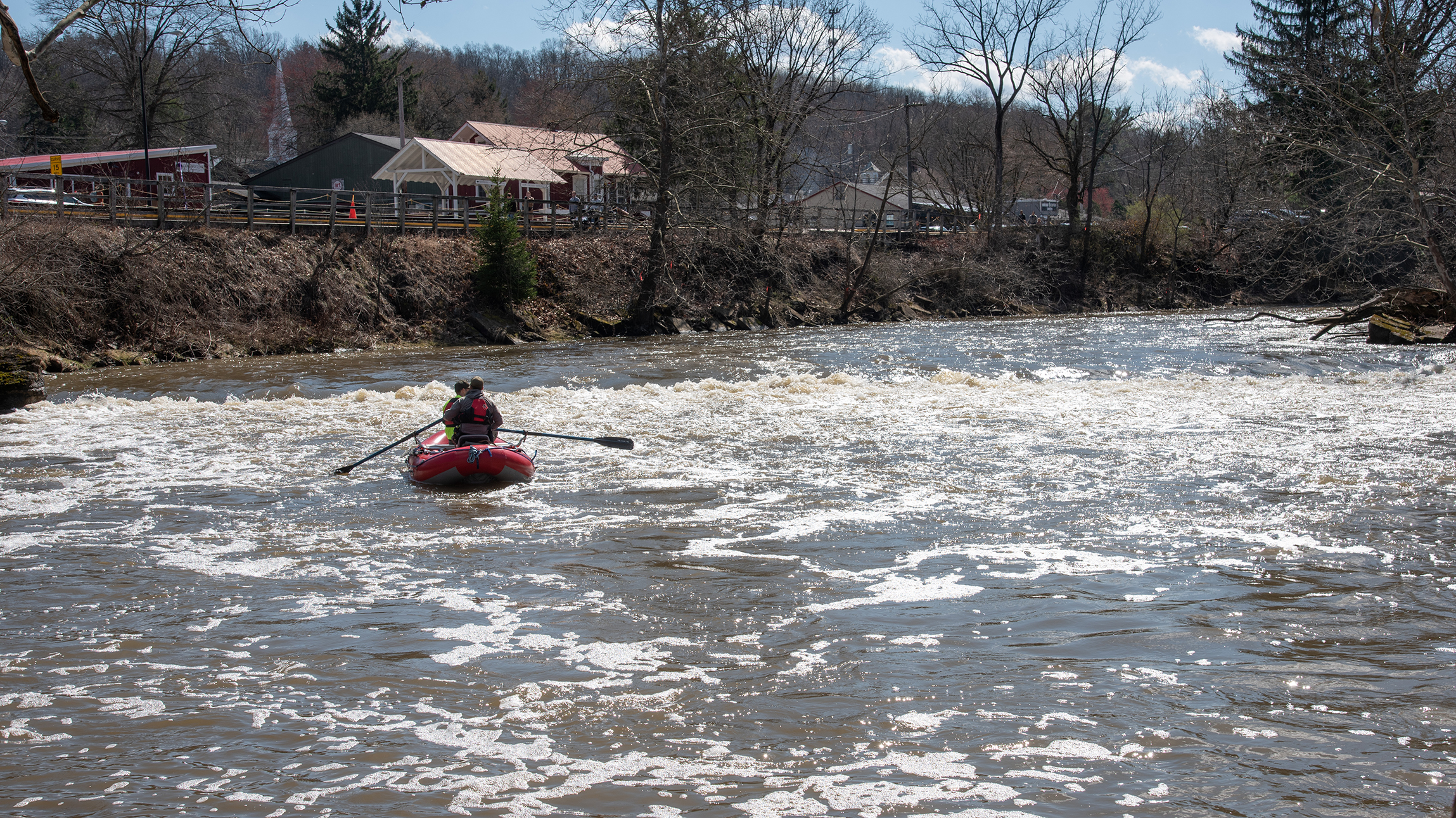 Two people paddle a red raft through riffles on a river; buildings and a wooden fence along the riverbank above.