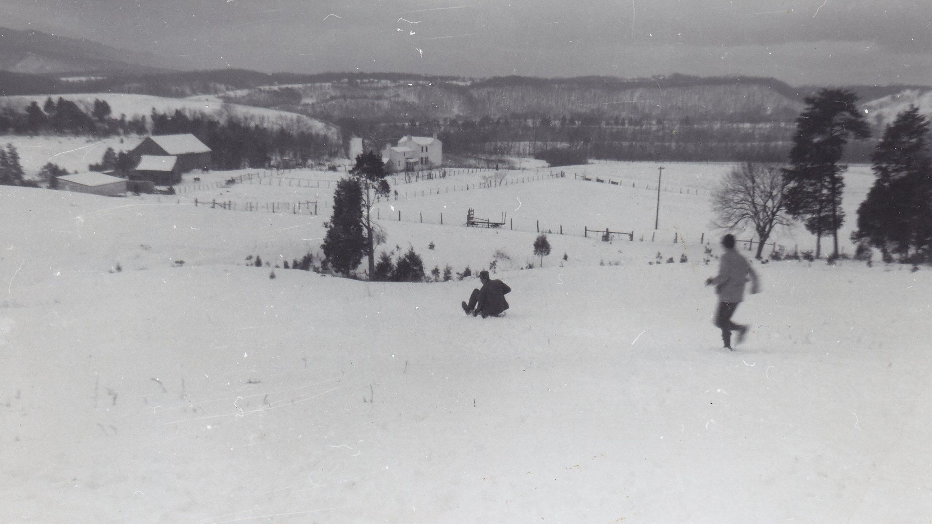 A landscape of downward sloping grassy fields. At the bottom of the hill is a two-story farm house and a large wooden barn.