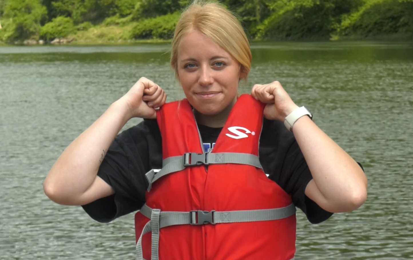Blond man wearing lifejacket. Lifejacket buckles are closed, but jacket fits very loosely. Man lifts jacket and the jacket's shoulders easily can be lifted over his ears.