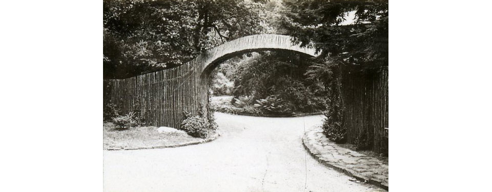 Black and white photo of entry arch spanning a driveway, connected to wooden fencing.