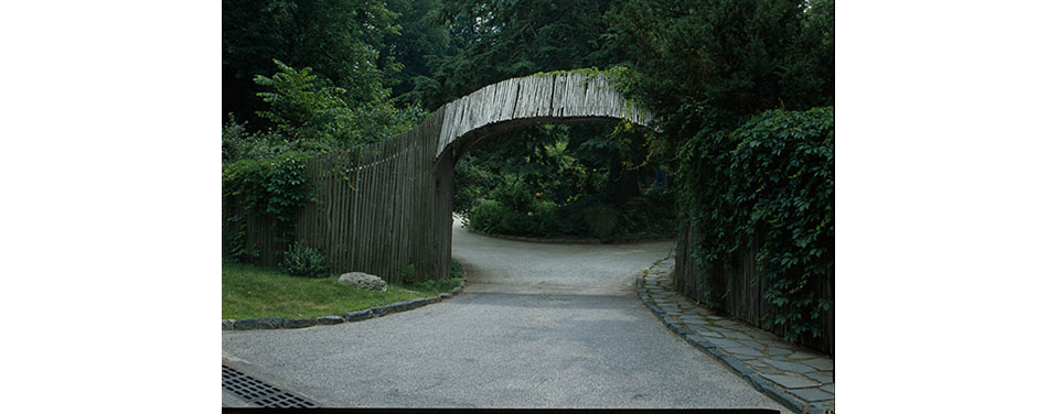 Black and white photo of entry arch spanning a driveway, connected to wooden fencing.
