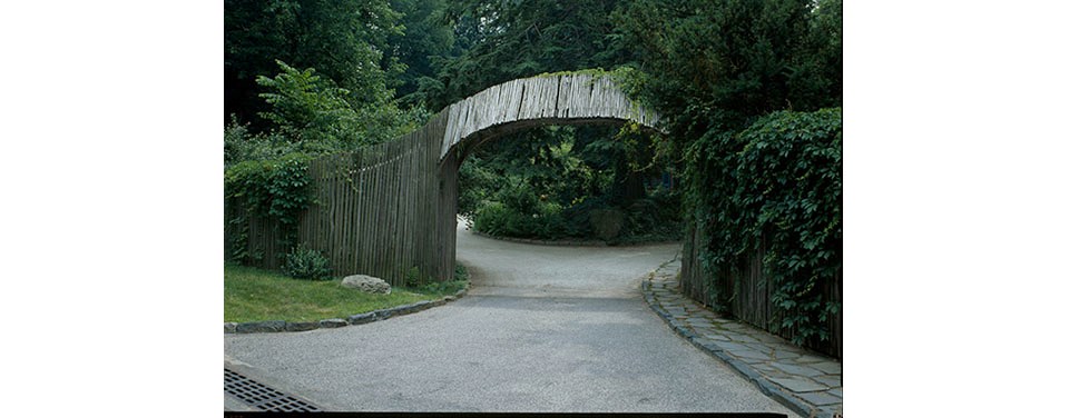 Black and white photo of entry arch spanning a driveway, connected to wooden fencing.