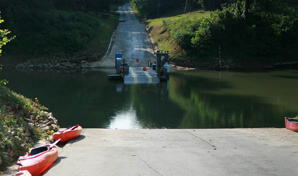 A person stands on wooden ferry at the end of a ramp, hands on hips. The road and concrete gutters continue on the other side of the water.