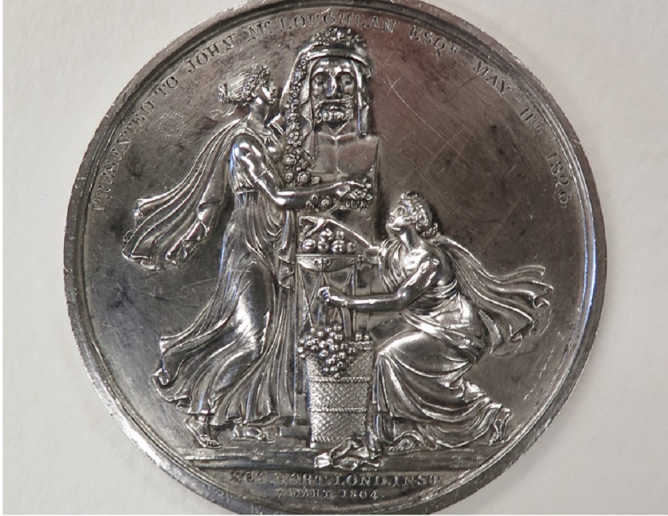 A dull silver medal showing two figures in flowing clothes kneeling on either side of a statue.