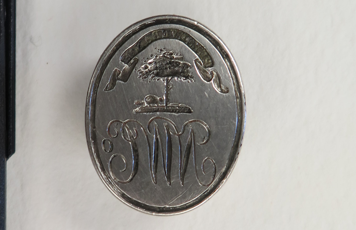 A metal seal with the letters NWC, a tree, and a banner with the word "Perseverance." The seal is darkened with age and grime.