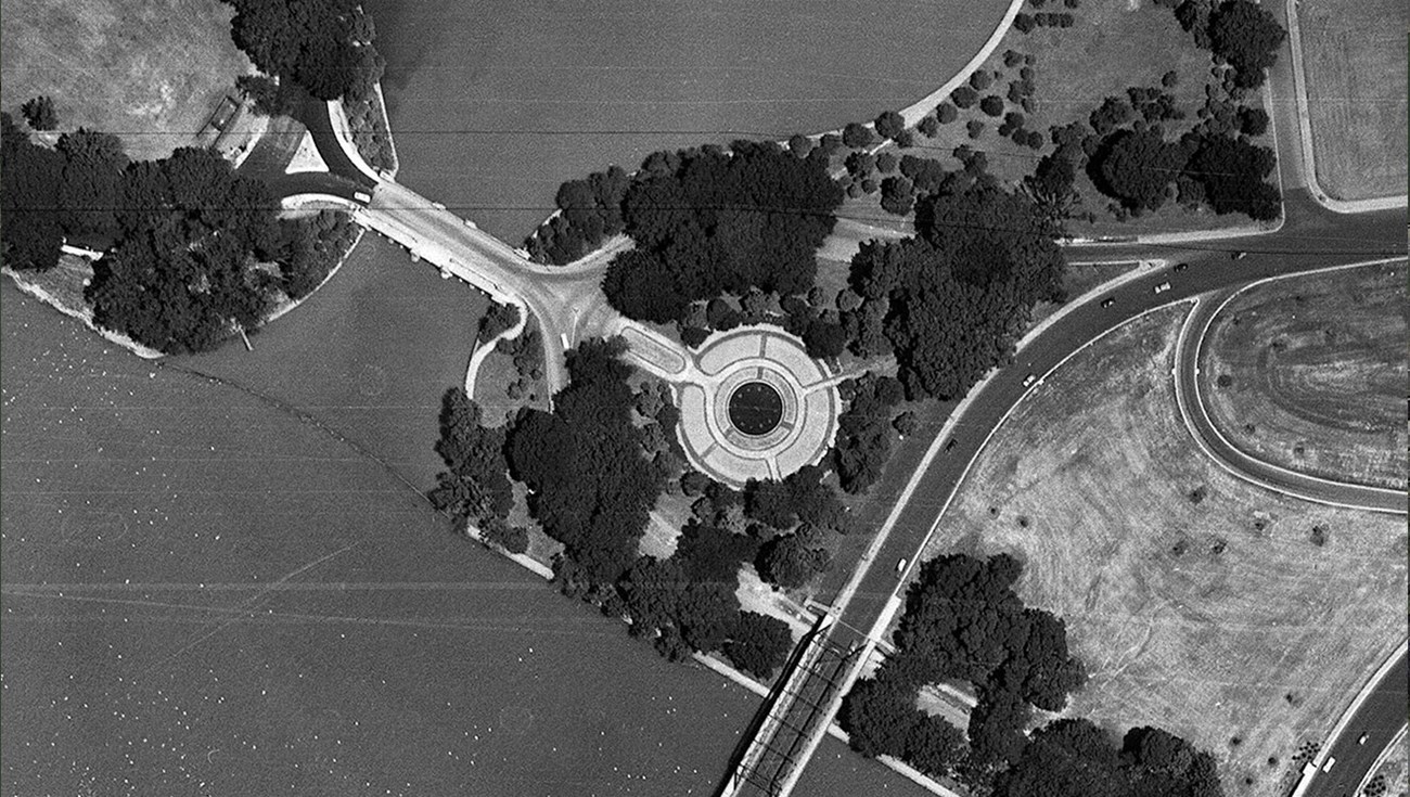 Aerial image of memorial landscape with round fountain and garden, thick canopy of trees, highway bridge over the river to the left, and cleared area to the right.