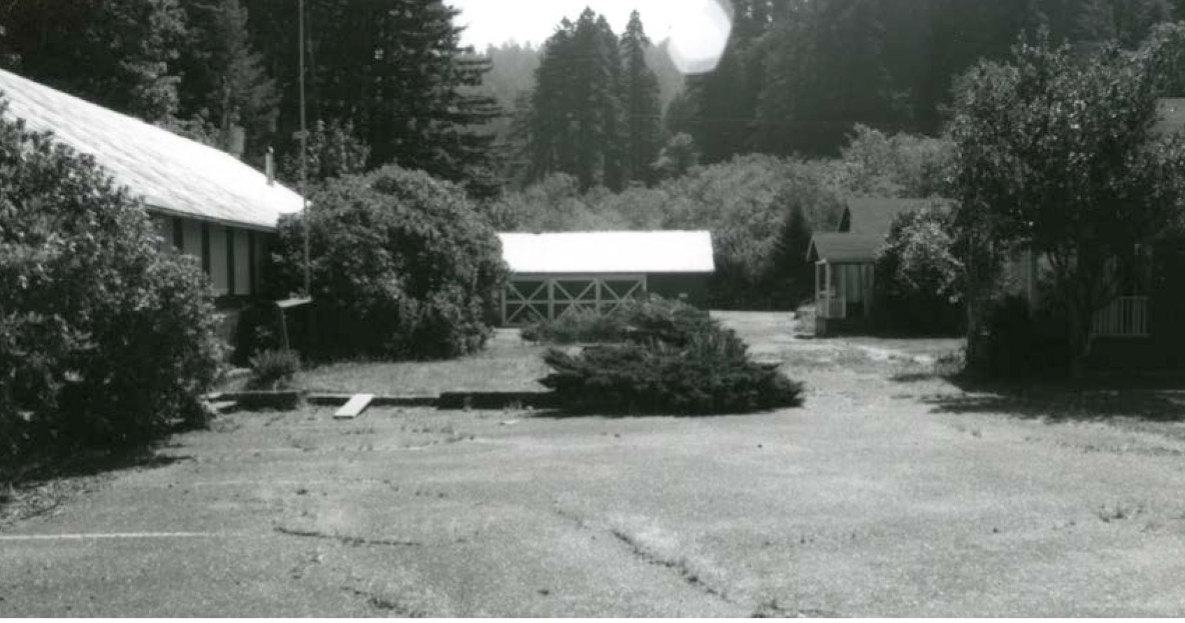 One-story buildings form a perimeter around an area of lawn and asphalt. Some shrubs grow at the foundations and a pond is in the center.