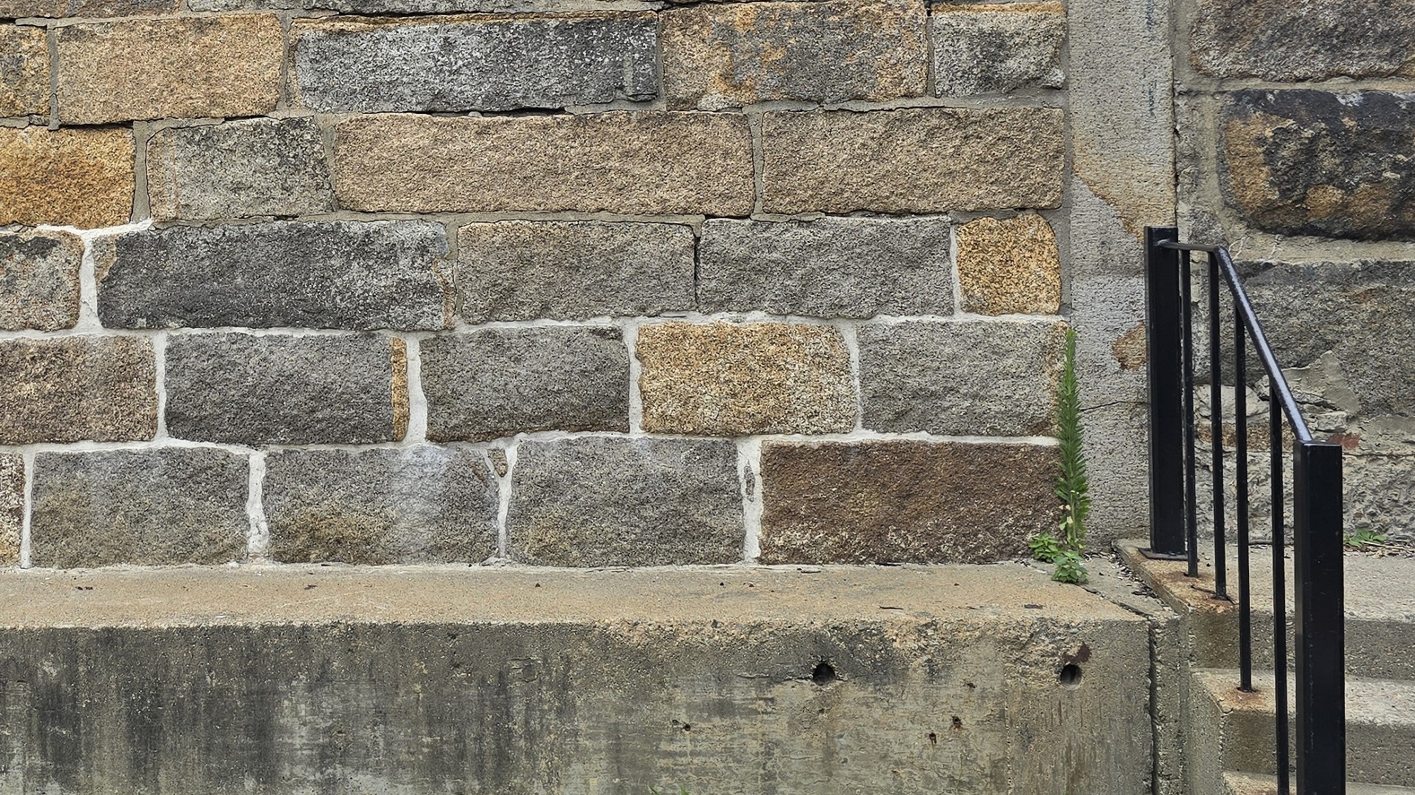 A closeup of a wall made from large, granite stone blocks. The mortar around the first two layers of blocks is newer and a brighter shade of grey than the mortar around the higher blocks.