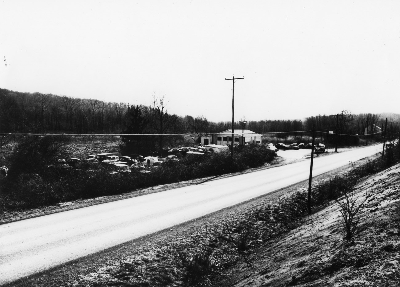 Black and white image looking across a two-lane road at a building surrounded by dozens of cars.