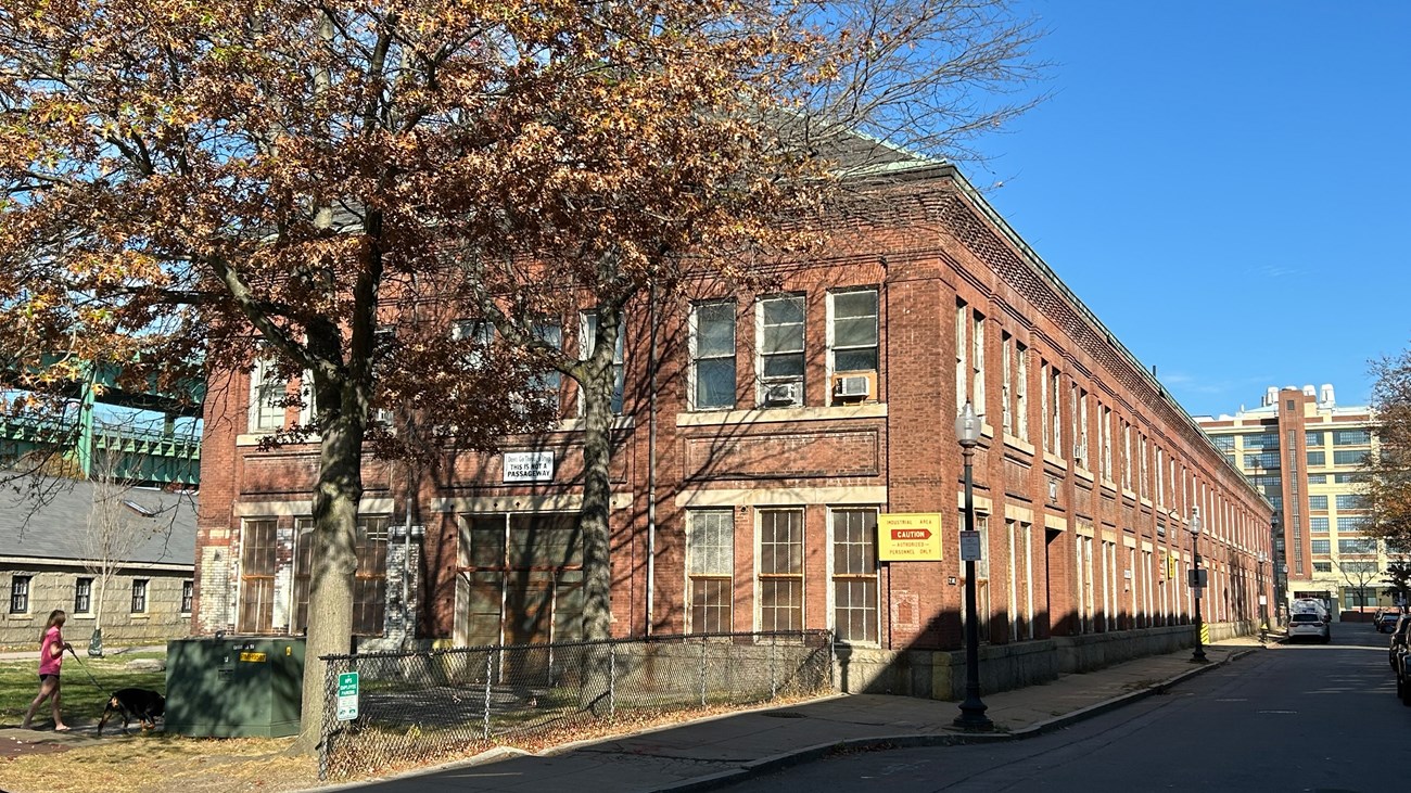 A long brick building that extends to the background. A tree in autumn sits before the building.