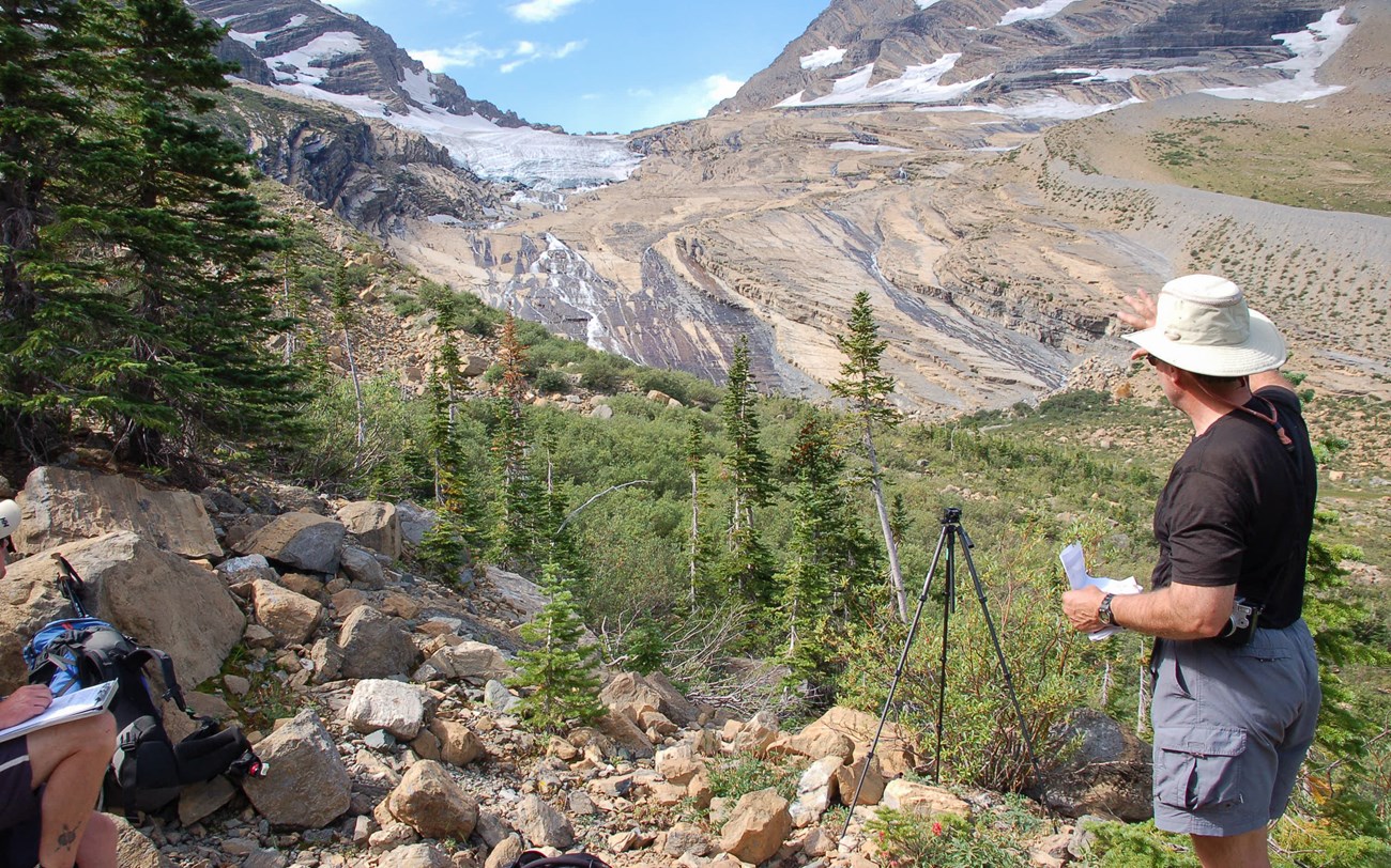 A person in a hat points at a mountain glacier on the right side of the image. Another person sits just outside the frame.