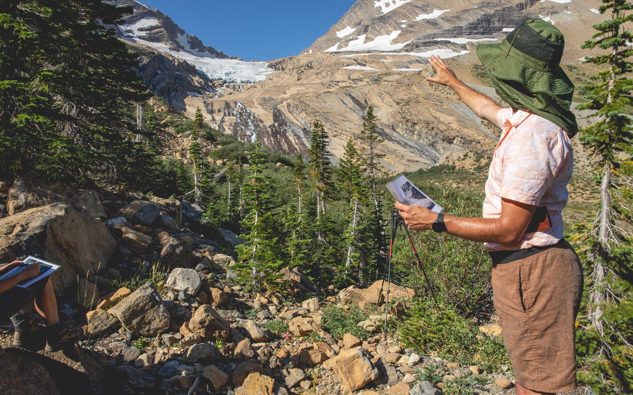 A person in a hat points at a mountain glacier on the right side of the image. Another person sits just outside the frame.