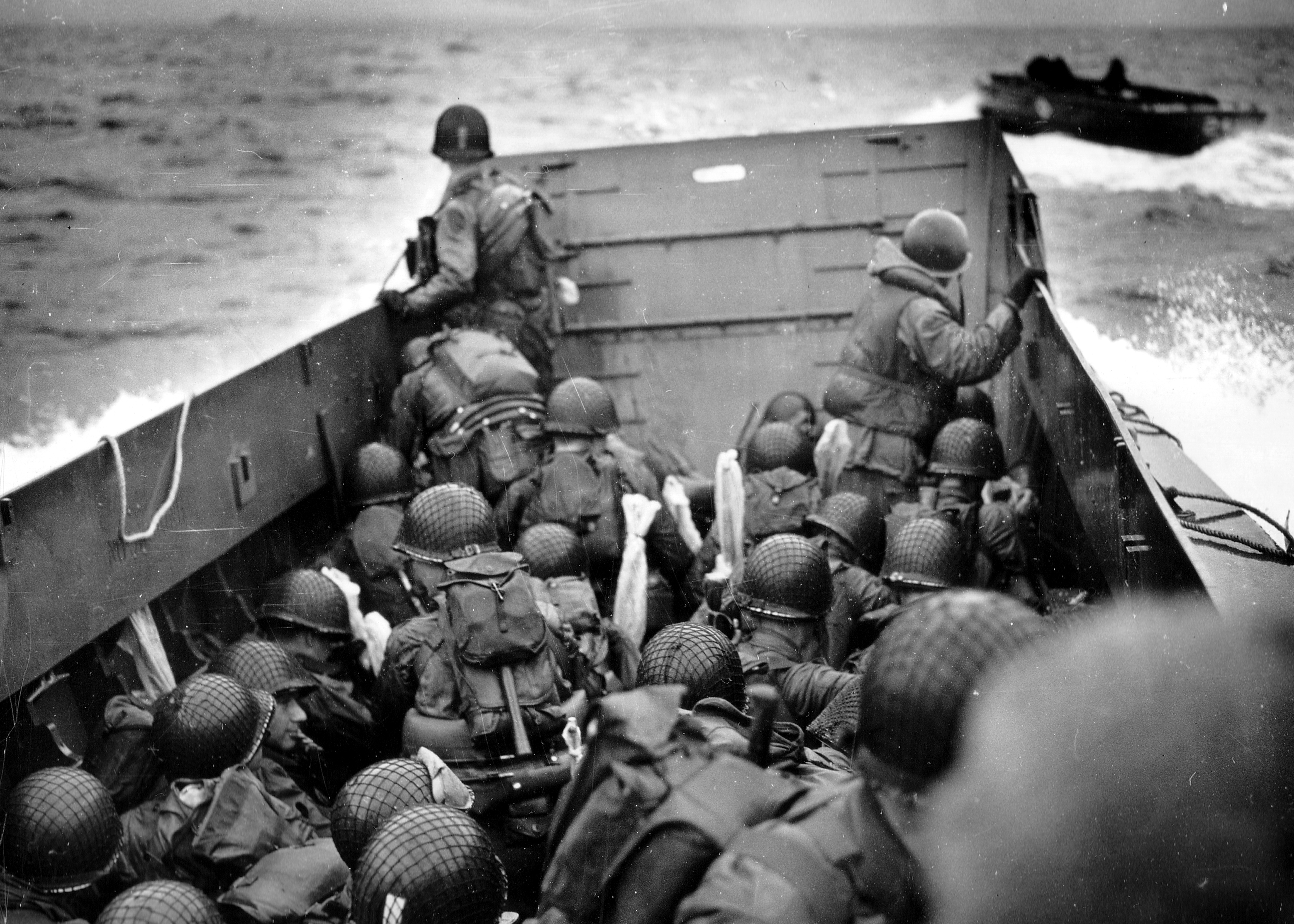 Troops in military gear crouch in a landing craft approaching a beach, exuding courage and tension. Ocean waves and another boat are visible.