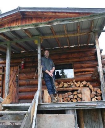 A man leans against a beam on the porch of a log cabin. Firewood sits on the porch behind him.