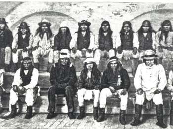 Black and white image of 16 Apache men sitting near a fort brick wall. 