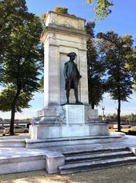 A monument featuring a dark bronze , standing figure atop an ornate stone base. Behind the figure is an ornate stone wall.