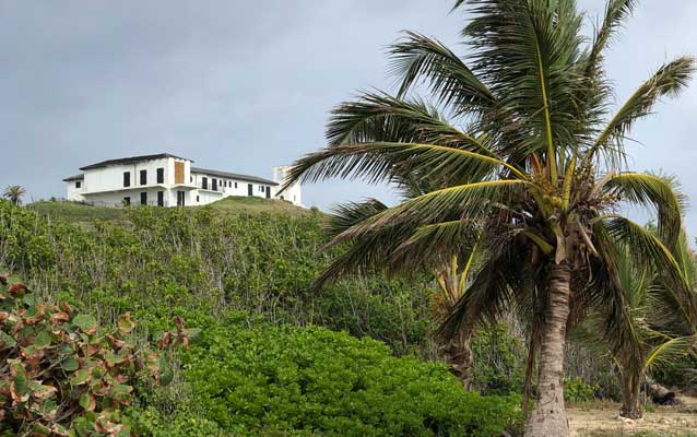 a white building up on cliff, beach and palm tree below