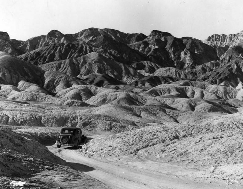 An old car drives around a curve with rocky mountains in background.