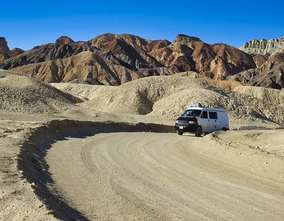 An old car drives around a curve with rocky mountains in background.