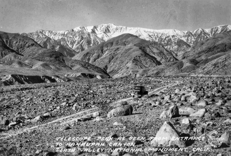 An old car drives on a dirt road with snow-capped peaks in background.