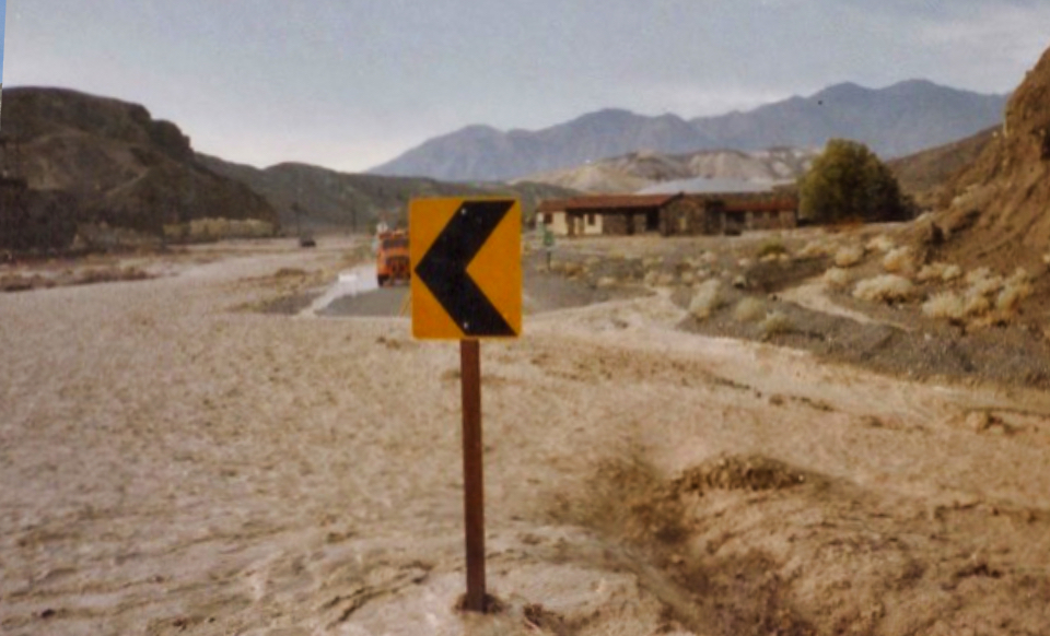 Rushing water covers road with buildings and mountains in background.