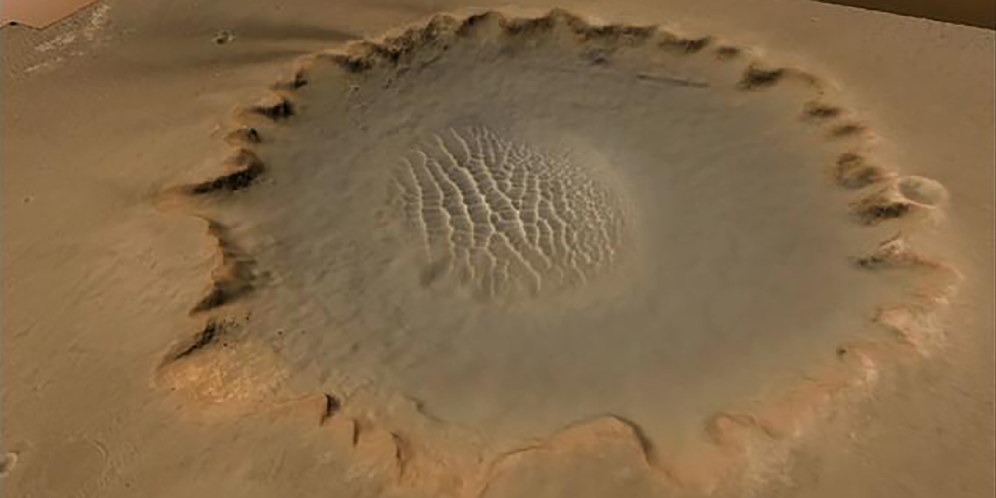 The photograph presents a vast, barren crater with steep, rocky sides and a pale, sandy bottom. It places emphasis on the crater's geological features, color stratifications, and visible man-made structures at its base.