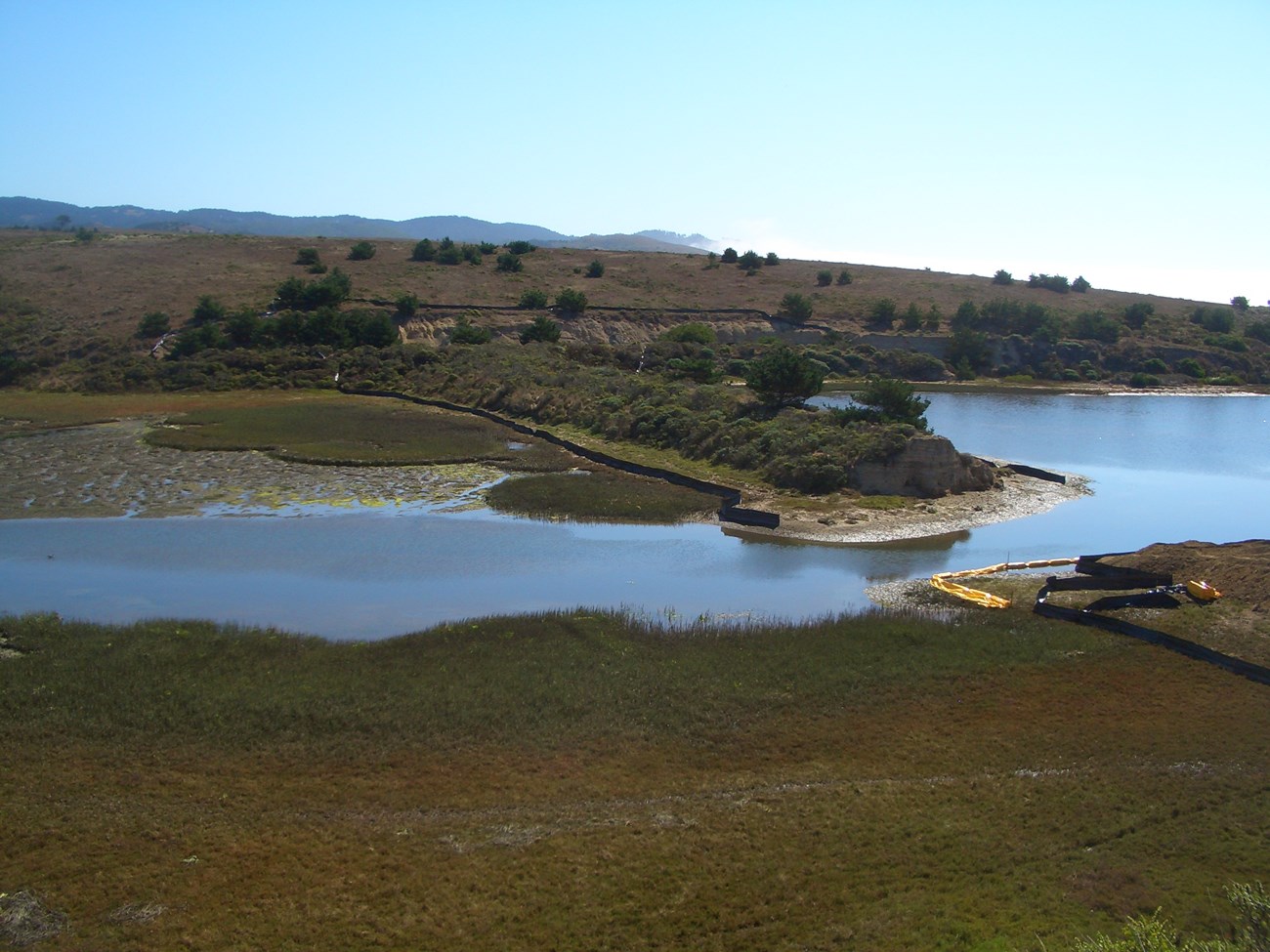 Photo of a very low-profile, shrub-covered dam that has been partially breached at the mouth of a shallow valley.