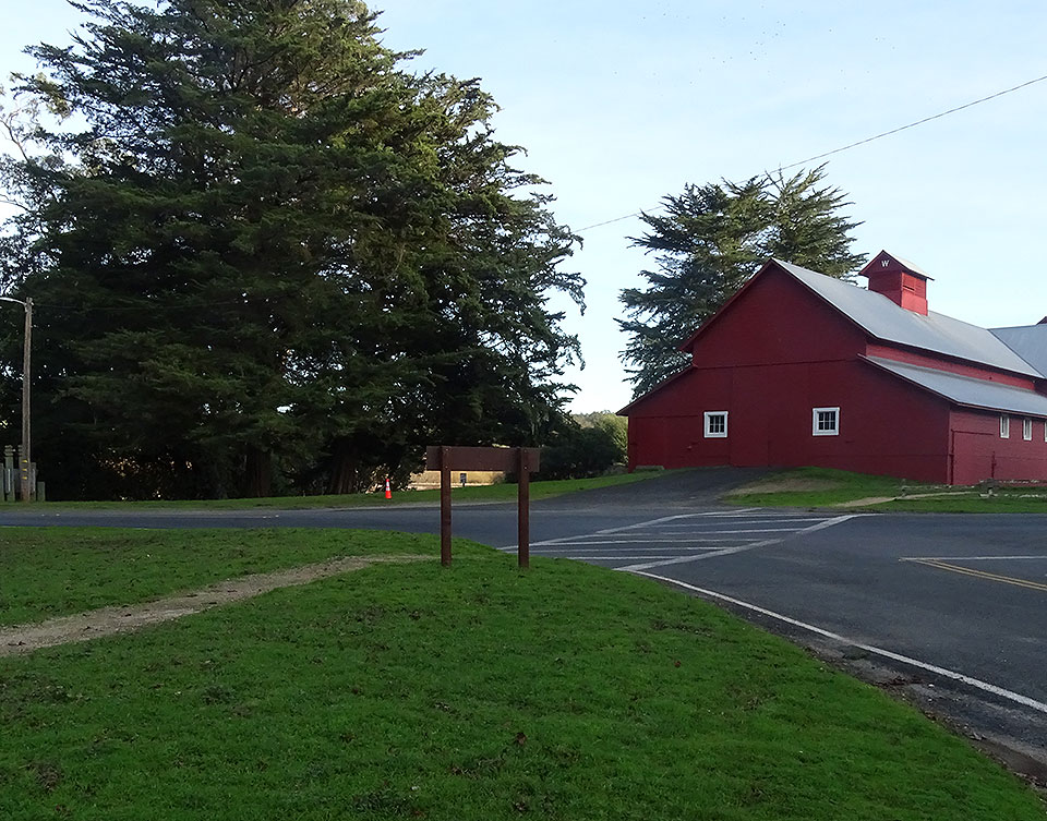 A black and white photo of 1960s automobiles parked to the right of a small building with an information sign and in front of an old barn.