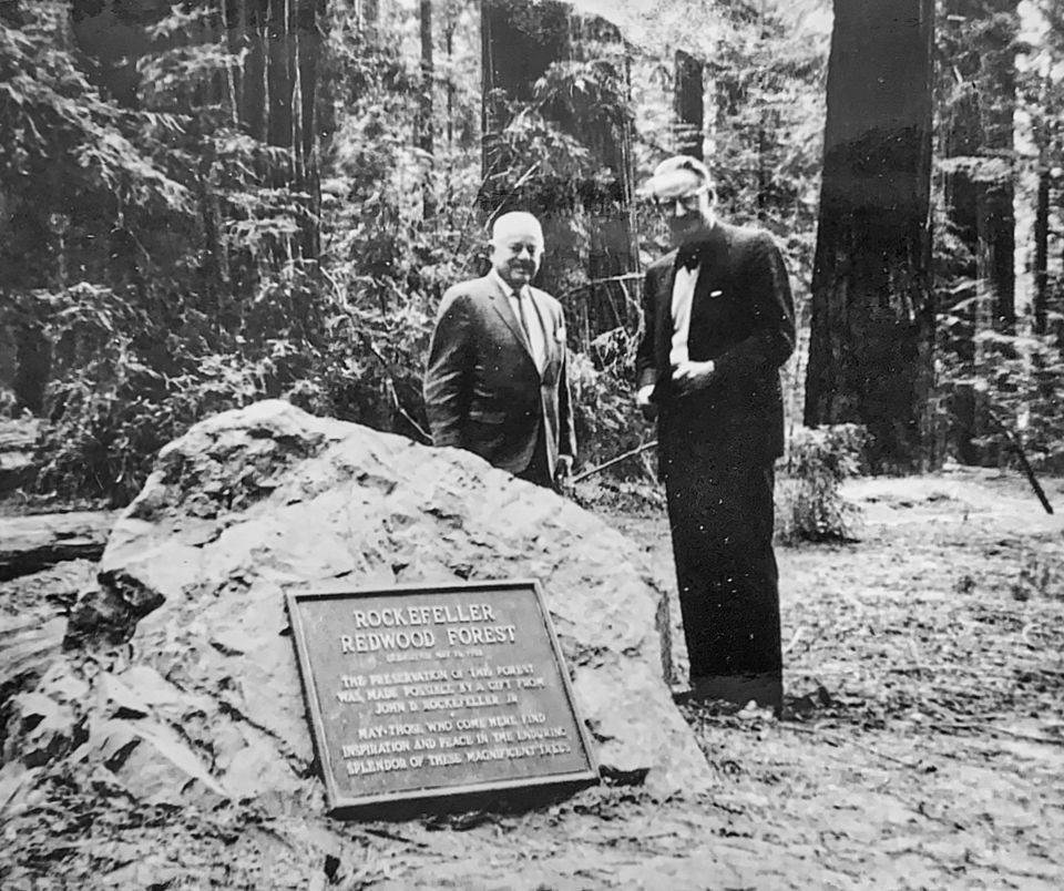 Two men stand behind rock with plaque, redwoods in background.